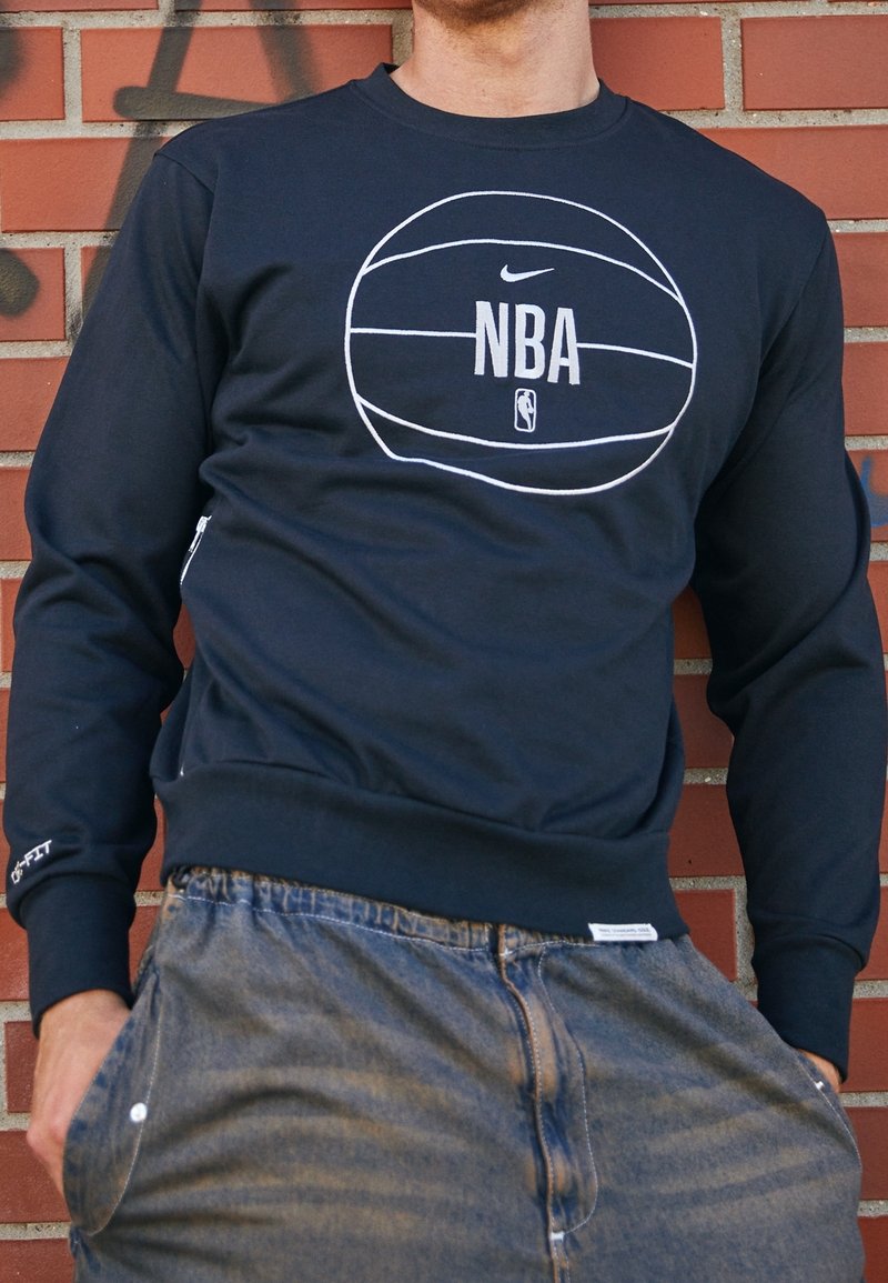 Homme portant un sweatshirt bleu marine avec un graphisme blanc de basketball NBA, debout contre un mur en briques rouges couvert de graffiti.