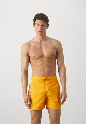Costumi da bagno gialli con vita regolabile, texture liscia e accenti bianchi a motivi sui lati. Il modello è senza maglietta.
