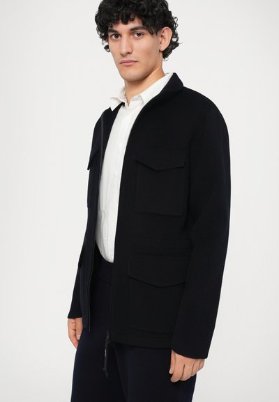 Tumesinine zip-up jakk, mis on valmistatud tekstuurilisest kangast, sisaldab kahte rinna taskut, küljetaskut ja kraega kaelust. Minimalistlik disain.