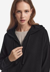 Czarna bluza z kapturem na zamek, wykonana z miękkiego materiału, z regulowanym kapturem. Model zdaje się trzymać zamek, odsłaniając beżowy inner top.