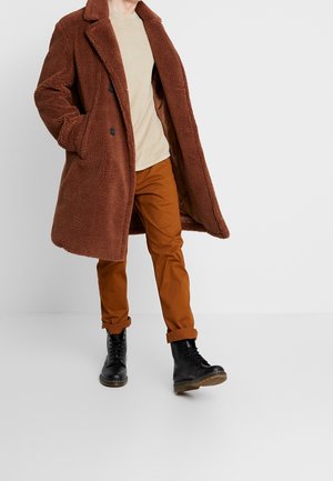 Hnědý ovčí kabát s klopovými límci, dvěma knoflíky, bočními kapsami, béžová košile, oranžové kalhoty a černé šněrovací boty.