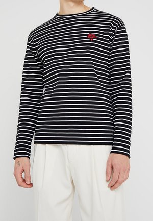 Chemise à rayures noires et blanches à manches longues avec un col rond et un détail brodé rouge sur la poitrine. Modèle portant un pantalon crème.