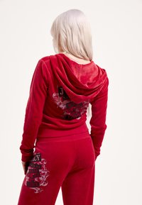 Piros velúr melegítő kapucnival. A pulóveren fekete és ezüst színű szöveggrafika található a háton. A nadrág oldalán ugyanolyan mintájú grafika van.