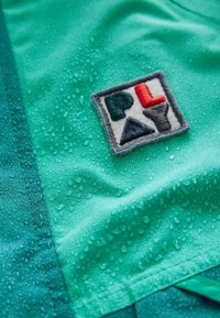 Grüner wasserdichter Stoff mit Regentropfen; verfügt über ein quadratisches Patch mit "PLAY" in roten, grünen und schwarzen Buchstaben, umrandet von grauem Stich.