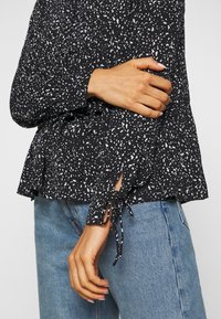 Blusa nera con motivo a macchie bianche, maniche arricciate e lacci ai polsini, indossata sopra jeans azzurri chiari.