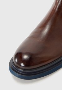 Bottine en cuir marron avec une finition brillante, bout arrondi et semelle en caoutchouc bleu. Présente un design sans couture et une forme générale élégante.
