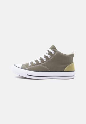 Zapatilla casual baja de lona verde oliva con cordones blancos, puntera de goma y suela blanca con rayas negras sobre un fondo neutro.