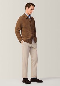 Giacca in suede marrone con tasche frontali, chiusura a bottoni; abbinata a pantaloni beige e scarpe slip-on in suede scuro. Il modello è in posa di profilo.