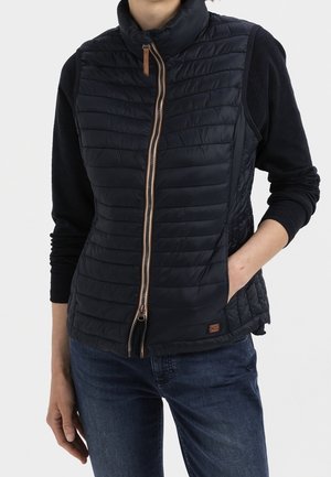 Personne portant une veste noire matelassée sans manches par-dessus une chemise noire à manches longues avec un jean bleu, main dans la poche de la veste, sur fond blanc.