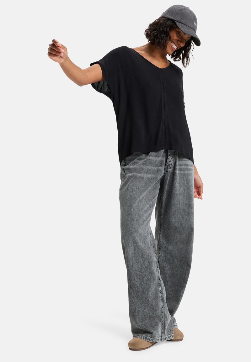 Un haut noir ample à manches courtes avec un col en V, associé à un jean gris taille haute à jambes larges et des mocassins beiges.