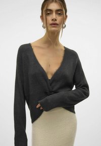 Cardigan tricoté gris foncé avec un décolleté en V profond et un nœud à l'avant ; manches longues et tissu texturé. Complété par une jupe légère.