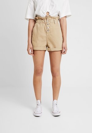 Vrouw draagt beige shorts met hoge taille, knoopsluiting aan de voorkant en opgerolde zoom, gecombineerd met een wit cropped topje en witte sneakers.