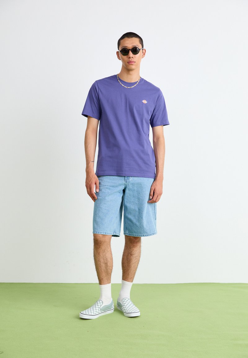 Lila Baumwoll-T-Shirt, Jeansshorts und karierte Slip-On-Schuhe. Accessoires sind eine runde Sonnenbrille und eine Kettenhalskette.