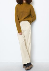 Personne debout contre un mur blanc portant un pull en maille moutarde, un pantalon large crème et des bottes à plateforme noires avec des semelles marron.