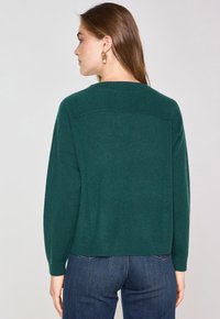 Maglione verde a maglia con una vestibilità morbida, collo rotondo e maniche lunghe. Texture liscia con polsini e fondo a coste, abbinato a jeans scuri.