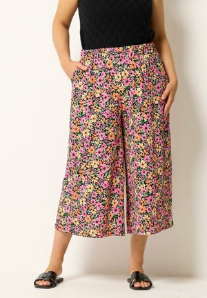 Kvinde iført vide bukser med blomsterprint i pink, orange og gul, sort ærmeløs top og sorte sandaler, hænderne i lommerne.