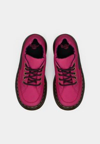 Pár jasne ružových topánok Dr. Martens s čiernymi šnúrkami, žltým prešívaním a hrubými hnedými podrážkami, pohľad zhora na bielom pozadí.