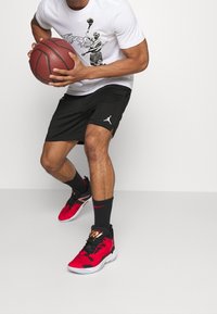 Black basketball shorts with mesh texture, featuring a white Jumpman logo. White T-shirt with a graphic print. Red and black basketball shoes.