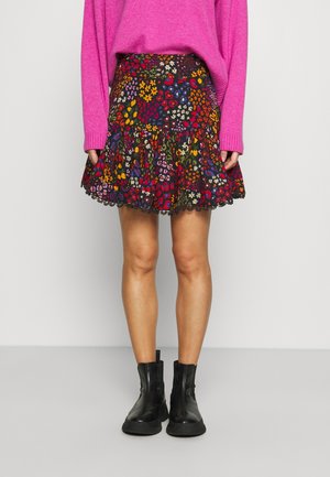 Kobieta ubrana w wielokolorową spódnicę w kwiaty i cętki pantery, różowy sweter oraz czarne botki, stojąca na tle jednolitego jasnego tła.