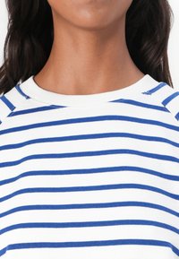 Camicia bianca con righe orizzontali blu, con scollatura tonda e maniche raglan. Tessuto morbido in misto cotone.