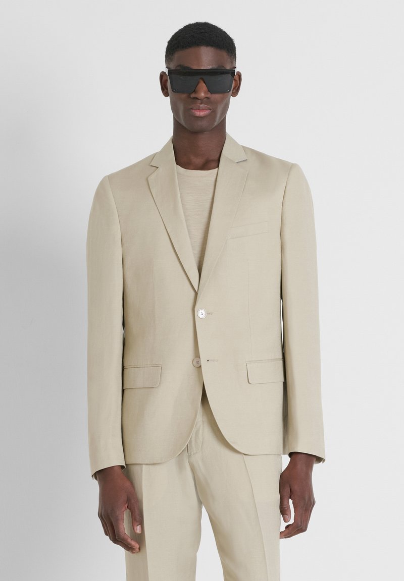 Costume en lin beige avec une coupe ajustée, deux boutons et des revers crantés. Assorti à une chemise coordonnée et de grandes lunettes de soleil noires.