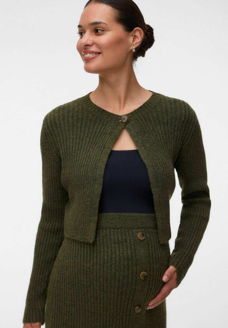 Femme aux cheveux foncés en chignon, portant un cardigan côtelé vert et une jupe assortie avec des boutons, souriante et tenant sa jupe d'une main.