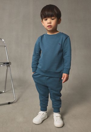 Niebieska bluza i dopasowane spodnie dresowe wykonane z miękkiego materiału, z małym logo. Stylizację dopełniają białe trampki.