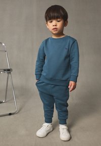 Sweat-shirt bleu et jogging assorti en tissu doux, avec un petit logo. Des baskets blanches complètent la tenue.