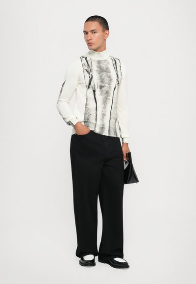 Λευκό πουλόβερ με ζιβάγκο που διαθέτει έναν μαύρο αφηρημένο σχεδιασμό, συνδυασμένο με μαύρα παντελόνια με φαρδιά γραμμή και μαύρα παπούτσια με λευκές λεπτομέρειες.