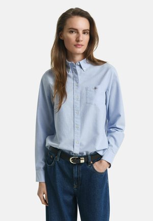 Young woman with long brown hair wearing a light blue button-up shirt tucked into dark blue jeans with a black belt, one hand in pocket.