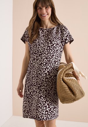 Lächelnde Frau mit langen braunen Haaren, die ein kurzärmeliges Kleid mit Leopardenmuster trägt und eine große geflochtene Strohtasche mit weißem Rand hält.