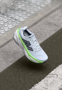 Világosszürke sneaker texturált felsőrésszel, fekete részletekkel és élénk zöld talppal. Jellemzője a kiemelkedő logó és a légáteresztő kialakítás.