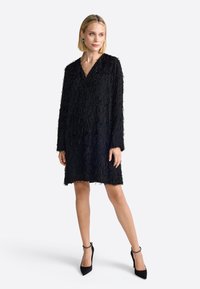 Fekete ruha textúrált bojt részletekkel, V-nyakkivágással, hosszú ujjakkal és laza fazonnal. A modellen fekete magassarkú cipők vannak.