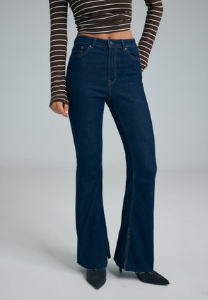 Femme portant un jean flare taille haute bleu foncé, associé à des bottes noires à bout pointu et un crop top rayé marron à manches longues.