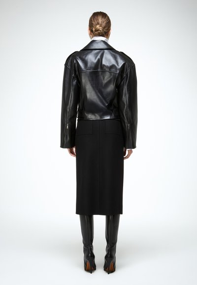 Osoba s kosom zavezanom unatrag, nosi crnu kožnu jaknu, crnu midi suknju i crne visoke čizme na petu do koljena, stojeći na bijeloj pozadini.