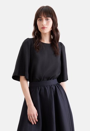 Kobieta o ciemnych, falowanych włosach nosi czarną sukienkę z szerokimi krótkimi rękawami i marszczoną spódnicą z wysokim stanem na jednolitym jasnym tle.