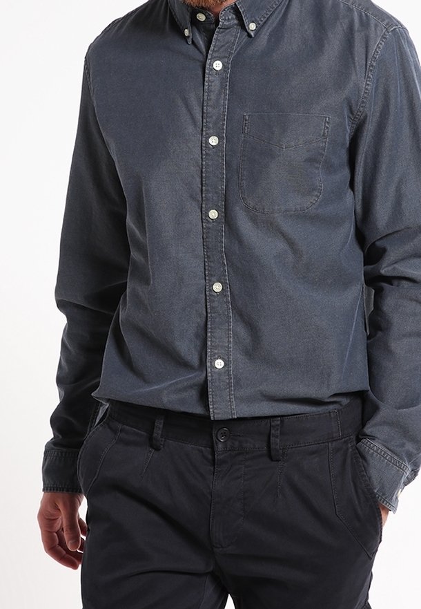 Chemise en denim foncé avec un col boutonné, une poche poitrine unique et des boutons argentés. Portée avec un pantalon foncé, mettant en avant un look décontracté.
