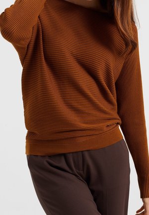 Persoon die een geribbelde, lange mouw, bruine trui draagt, ingestopt in donkerbruine broek, tegen een effen lichte achtergrond.