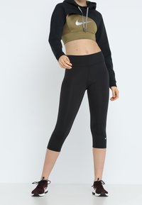 Felpa nera corta con un reggiseno sportivo verde oliva sotto, abbinata a leggings a cappri neri e scarpe da ginnastica rosso scuro.