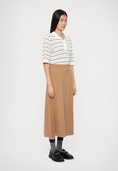 Donna in piedi di lato, vestita con una maglietta a righe bianca a maniche corte, una gonna midi color cammello, calze grigie e mocassini neri.