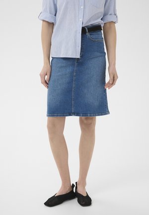 Femme portant une chemise bleu clair à manches retroussées, une jupe en jean bleu jusqu’aux genoux avec une ceinture noire, et des chaussures noires en mesh à enfiler, debout.