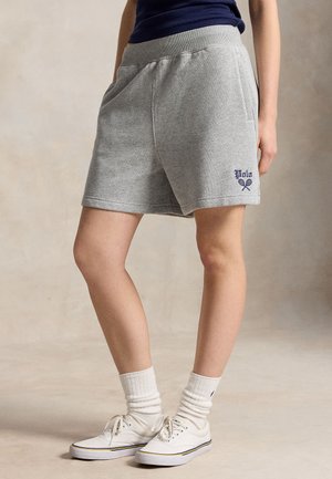 Osoba w szarych szortach z logo Polo, białych skarpetkach i białych trampkach, stojąca na neutralnym tle.