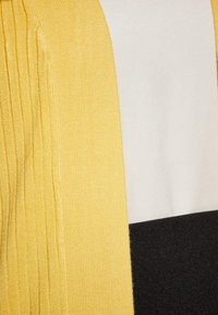 Sárga kötött anyag ribbed textúrával, sima fehér és fekete panelek kíséretében. A design három szakaszban színblokkoló elemeket tartalmaz.