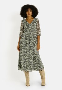 Vestido maxi de tela ligera y transparente con un estampado de serpiente en verde y negro. Escote en V y mangas abullonadas, combinado con botas beige.