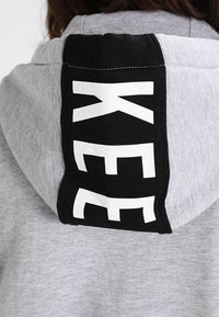 Grauer Hoodie mit einer schwarzen Rückenpartie, die auffälligen weißen Text zeigt. Das Material wirkt weich, mit glatter Textur und regulärer Passform.