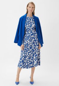 Blauer Blazer über einem weißen und blauen gemusterten Kleid. Das Kleid weist abstrakte Formen auf. Das Model trägt blaue spitze Absätze und silberne Ohrringe.