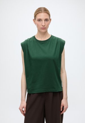 Femme à la peau claire et aux cheveux blonds attachés porte un haut sans manches vert foncé et un pantalon large marron foncé, debout devant un fond clair uni.