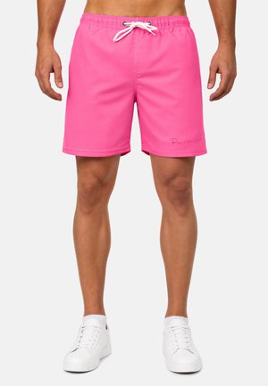 Homme portant un short de bain rose vif avec un cordon blanc et des baskets blanches, debout devant un fond uni.