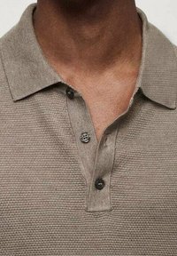 Gros plan sur une personne portant un polo taupe texturé avec un col large et trois boutons foncés, deux boutonnés et un déboutonné.