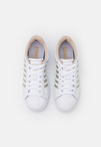 Bílé tenisky se třemi metalickými zlatými pruhy, šněrovacím designem a polstrovaným límcem. Texturovaná gumová podrážka, hladký kožený svršek.